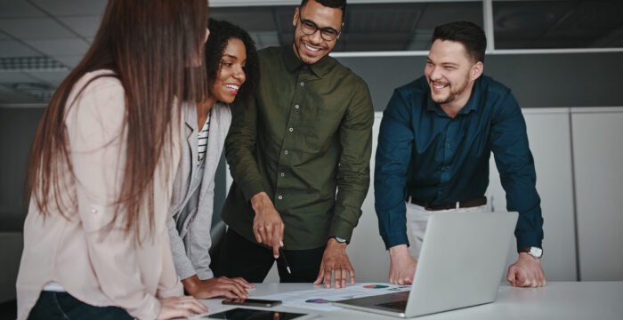 Group of young multiracial business people standing together at the office desk discussing business strategy looking at laptop Cheerful diverse business team working on project with laptop in office